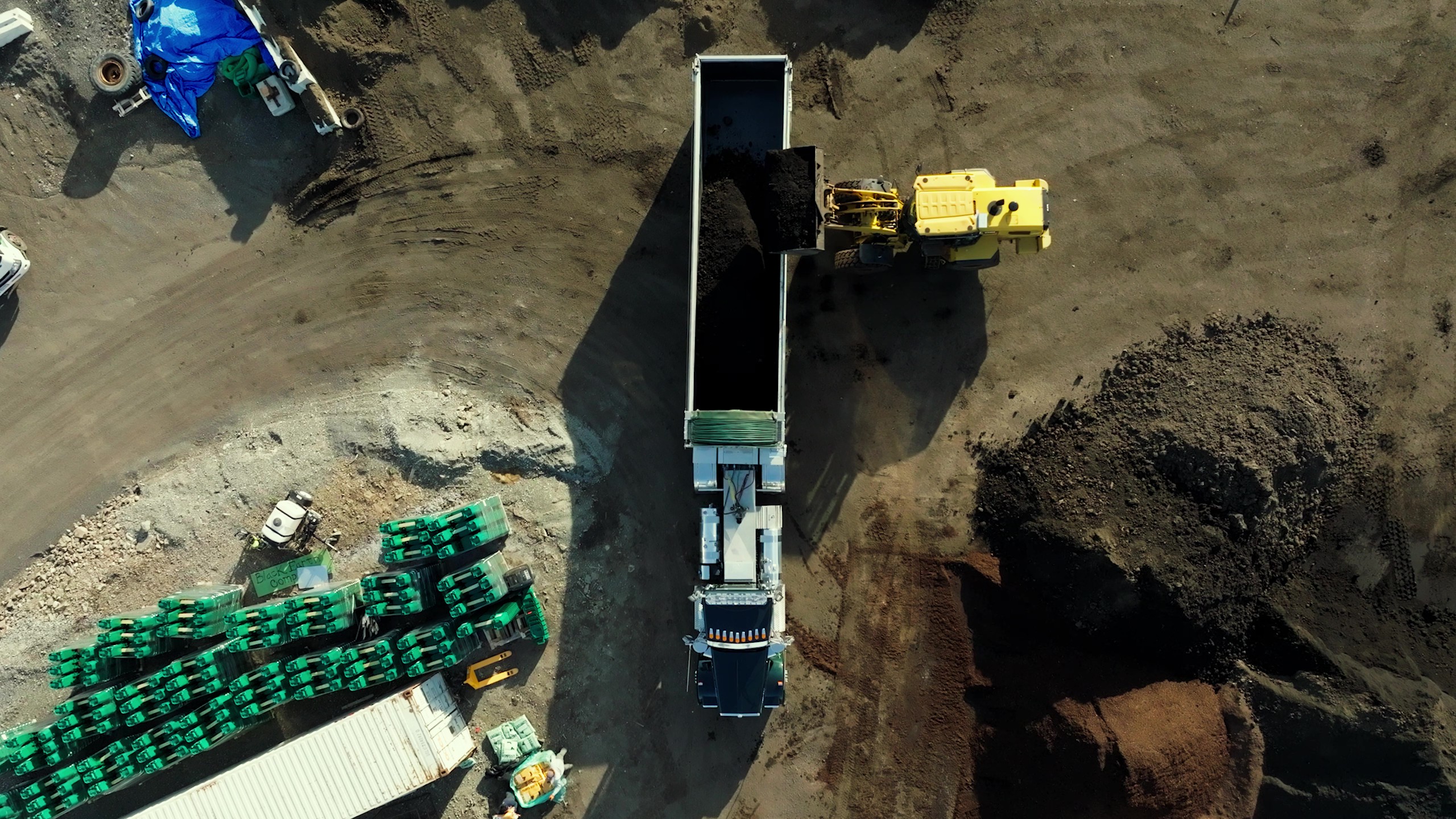 Aerial view of a yellow loader dumping soil into a dump truck at a construction site with green pallets and piles of soil nearby.