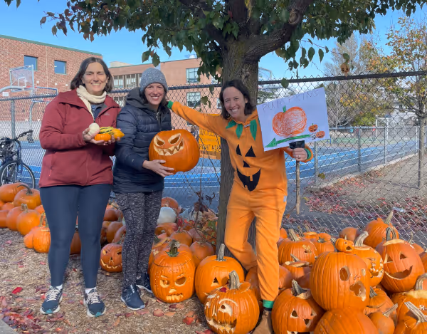 Three women standing outdoors near a tree and a display of carved pumpkins, one wearing a jack-o'-lantern costume holding a pumpkin drawing.