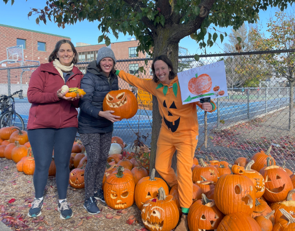 Three women standing outdoors near a tree and a display of carved pumpkins, one wearing a jack-o'-lantern costume holding a pumpkin drawing.