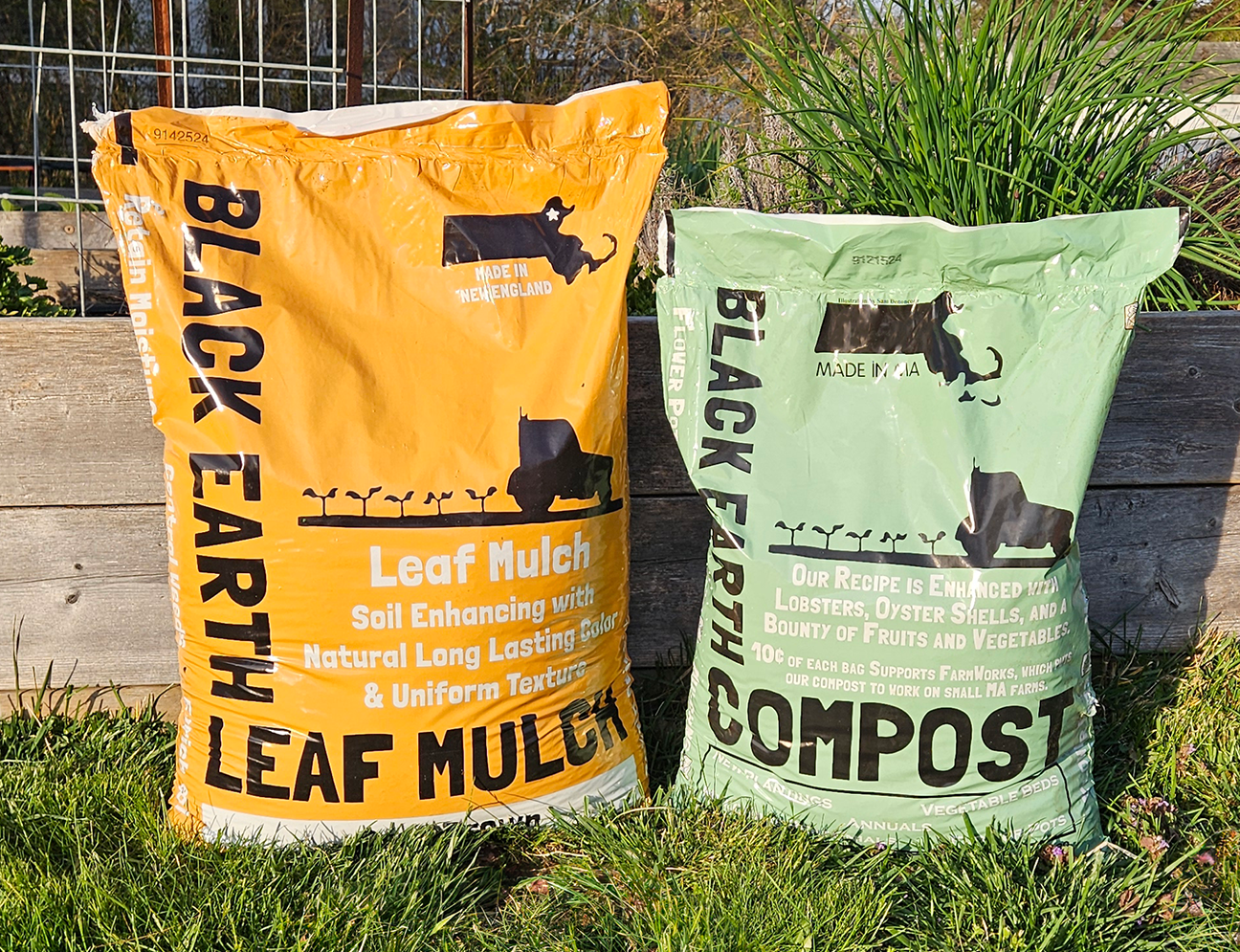 Two bags of Black Earth brand gardening products on grass; one orange bag labeled Leaf Mulch and one green bag labeled Compost.