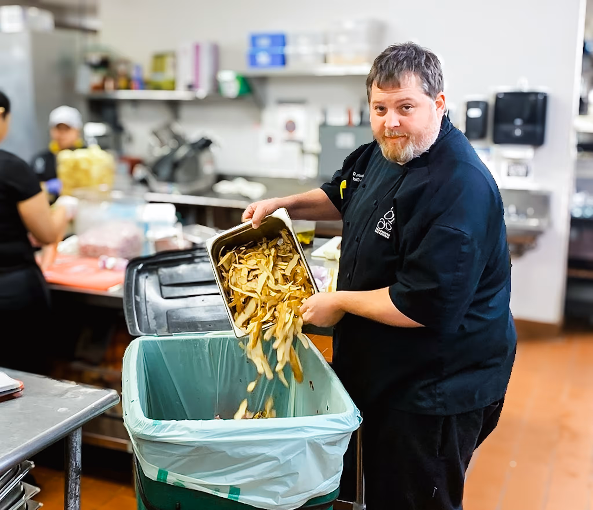 Chef in a kitchen emptying a tray of potato peelings into a large green compost bin.