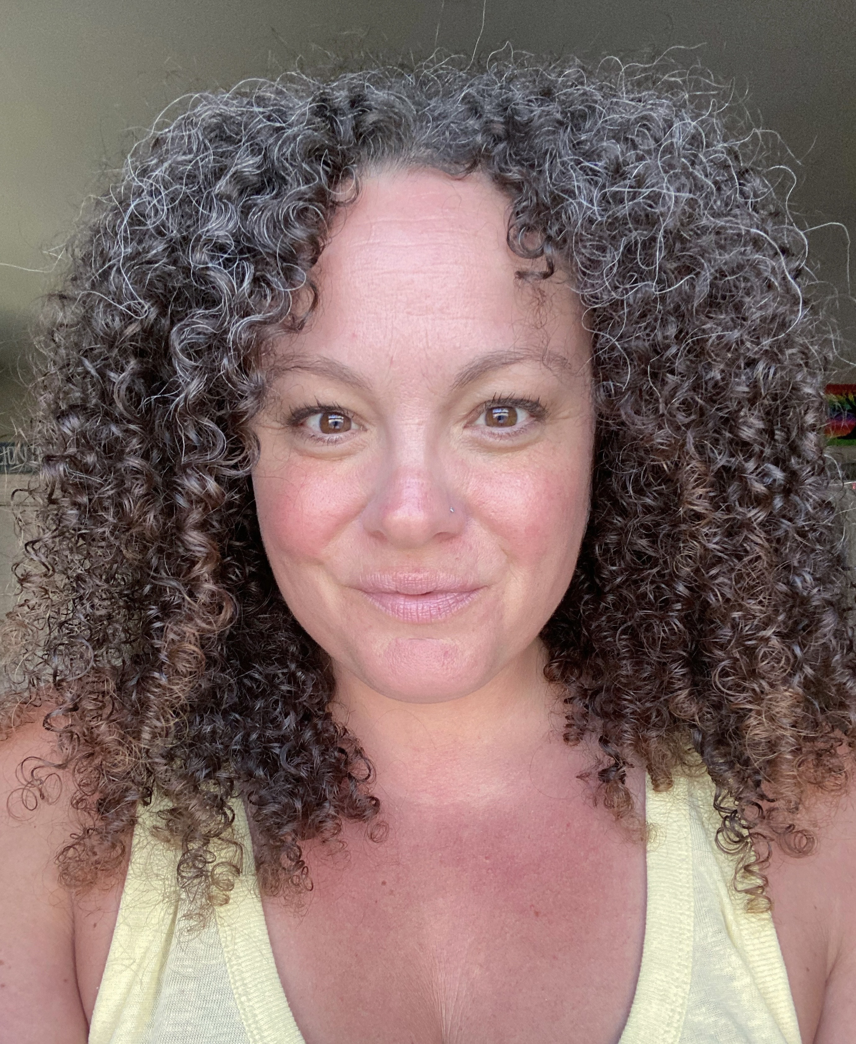 Close-up of a woman with curly brown and gray hair wearing a yellow tank top, smiling slightly at the camera.