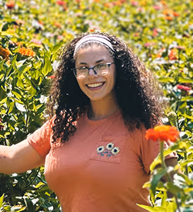 Smiling woman with curly hair and glasses standing among vibrant green plants and flowers outdoors.