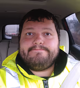 Bearded man wearing a neon yellow jacket sitting in a car with a seatbelt on.