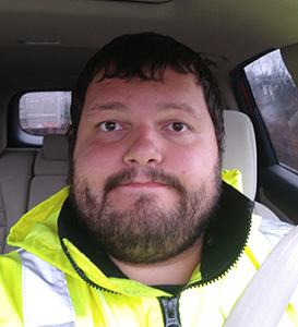 Bearded man wearing a neon yellow jacket sitting in a car with a seatbelt on.