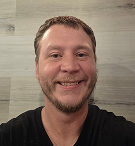 Smiling man with short brown hair and beard wearing a black shirt in front of a light gray wooden wall.