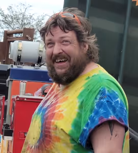 Smiling man with beard and curly hair wearing a colorful tie-dye shirt and orange sunglasses on his head outdoors.
