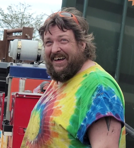 Smiling man with beard and curly hair wearing a colorful tie-dye shirt and orange sunglasses on his head outdoors.