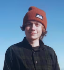 Young person wearing an orange beanie and a dark plaid shirt smiling against a clear blue sky.