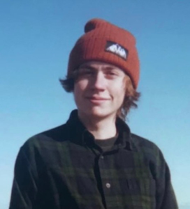 Young person wearing an orange beanie and a dark plaid shirt smiling against a clear blue sky.