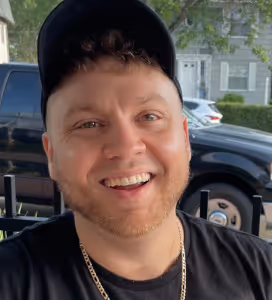 Smiling man with short curly hair, beard, black cap, and gold chain, sitting outdoors in front of parked cars and houses.