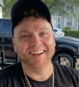 Smiling man with short curly hair, beard, black cap, and gold chain, sitting outdoors in front of parked cars and houses.
