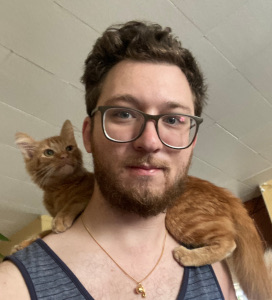 Man wearing glasses and a tank top with a ginger cat resting on his shoulder.
