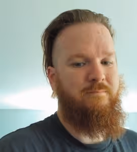 Man with a red beard and mustache, light skin, short dark hair on top and shaved sides, wearing a dark shirt, against a light blue background.