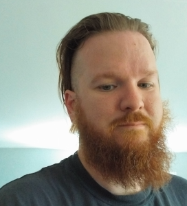 Man with a red beard and mustache, light skin, short dark hair on top and shaved sides, wearing a dark shirt, against a light blue background.