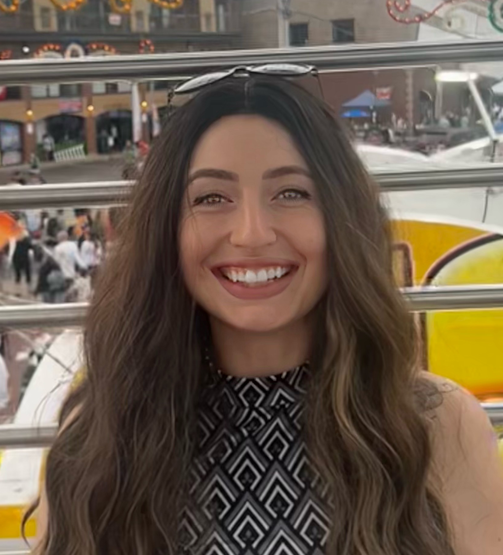 Smiling woman with long wavy brown hair, wearing a black and white geometric patterned top and sunglasses on her head, at an outdoor location with people and buildings in the background.