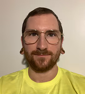 Man with short brown hair, beard, glasses, large ear gauges, and wearing a yellow shirt against a plain light wall.