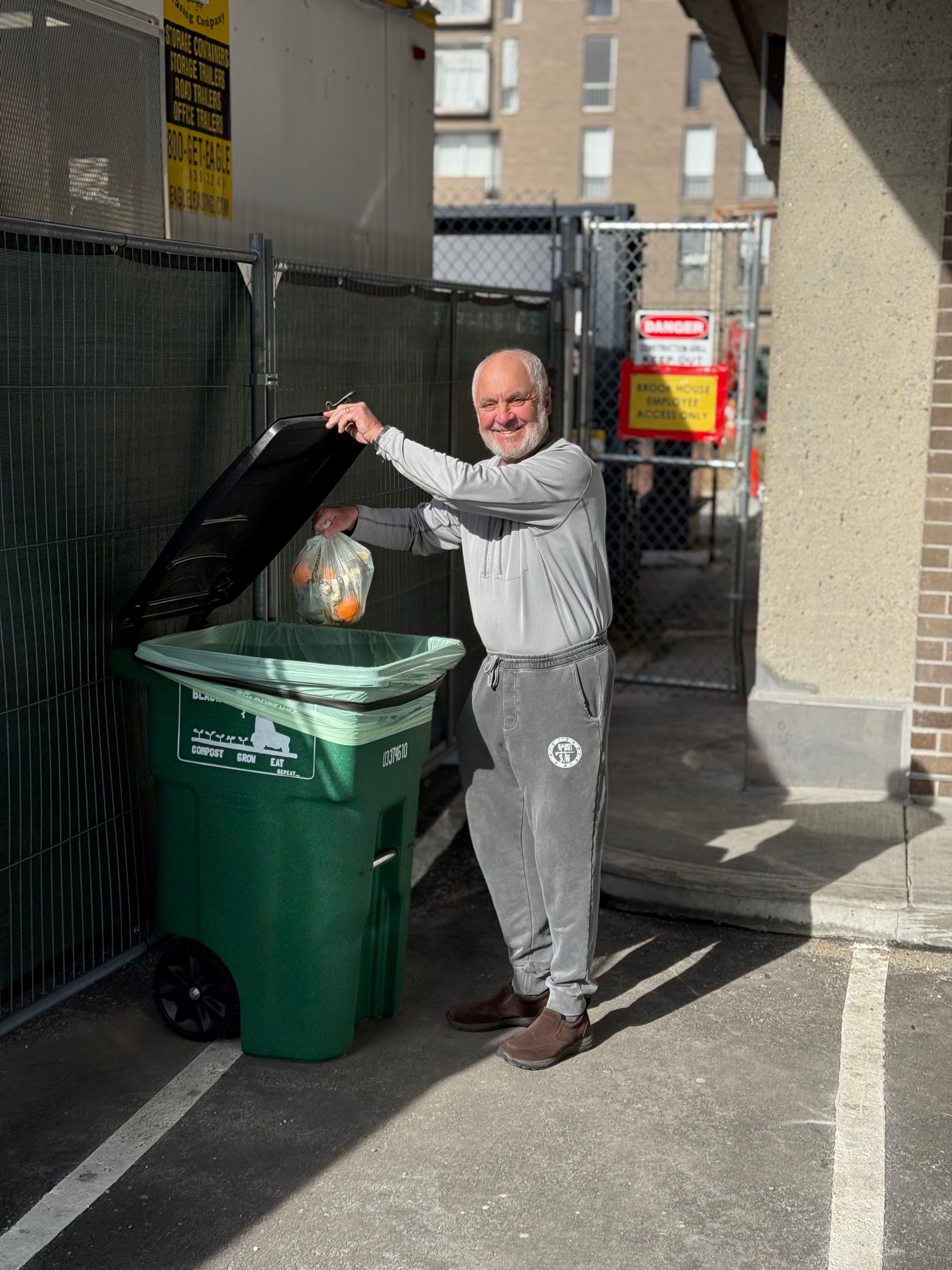Smiling elderly man throwing a bag of compostable food waste into a green compost bin outside.