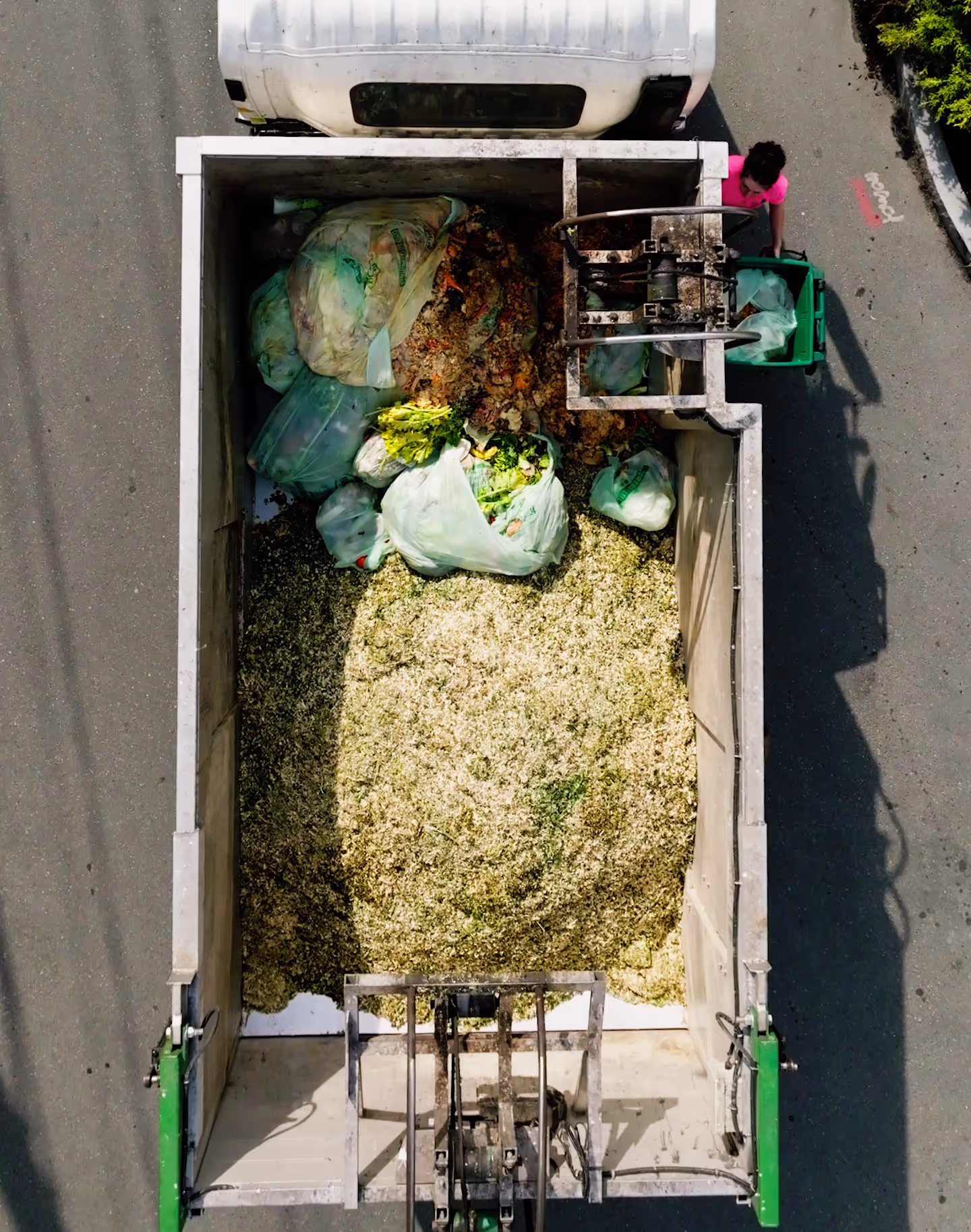 Top aerial view of a garbage truck filled with organic waste bags and food scraps, with a worker in a pink shirt placing waste into the truck.