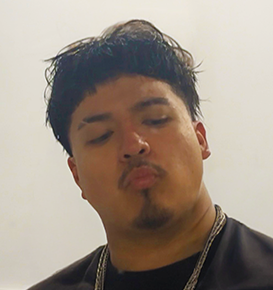 Portrait of a man with short black hair, a mustache, and goatee, wearing a dark shirt and chains, against a plain light background.