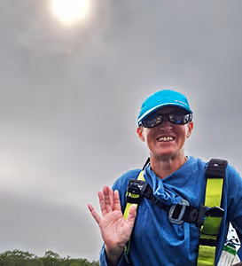 Person wearing sunglasses and a blue cap, waving and smiling outdoors under a bright sun with cloudy sky.