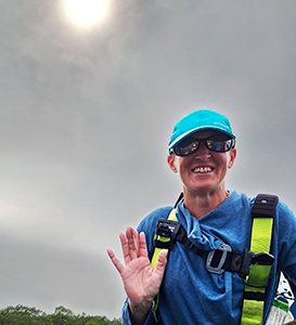 Person wearing sunglasses and a blue cap, waving and smiling outdoors under a bright sun with cloudy sky.