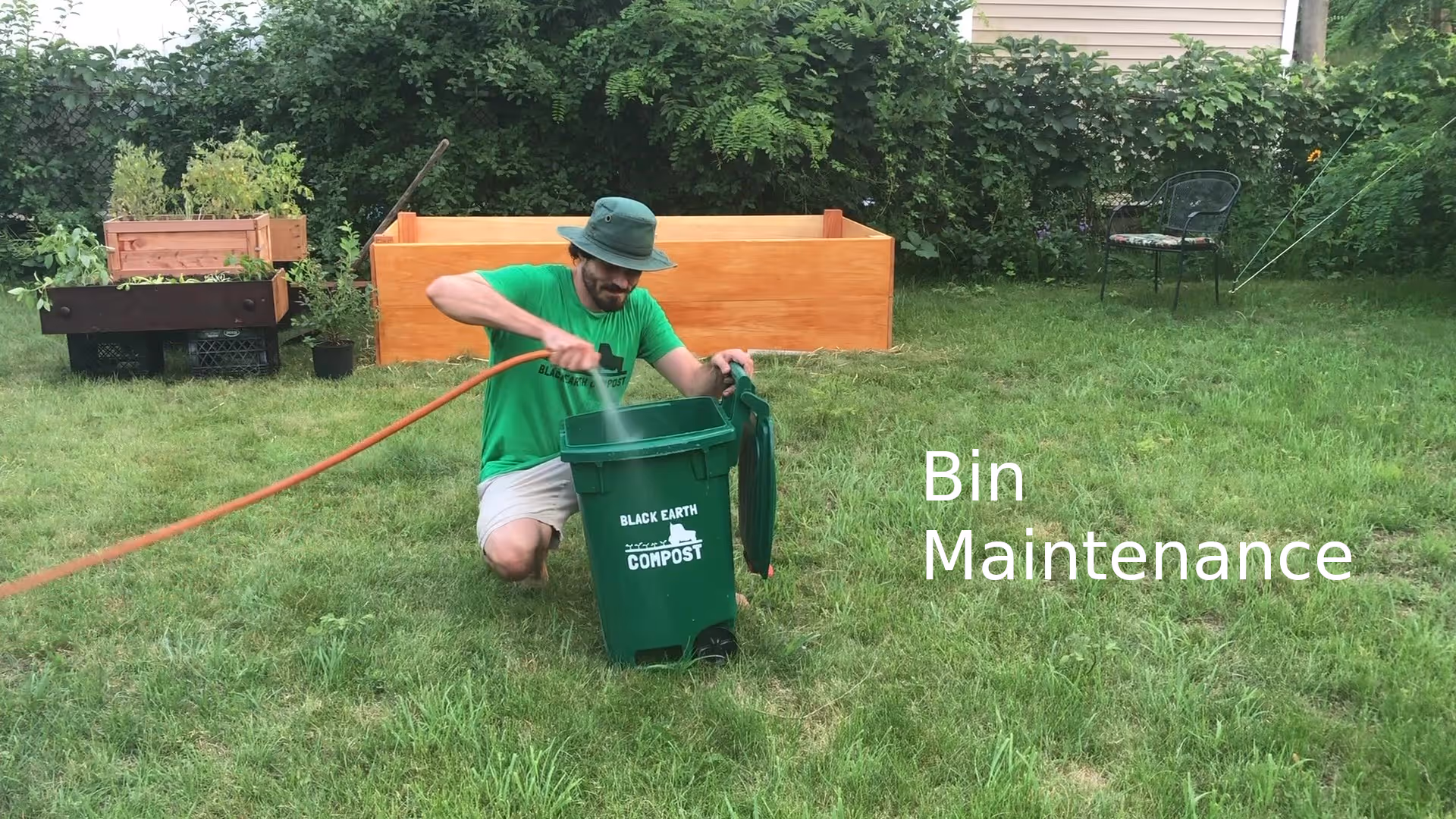 Man in green shirt and hat watering a green compost bin labeled Black Earth Compost in a grassy backyard.