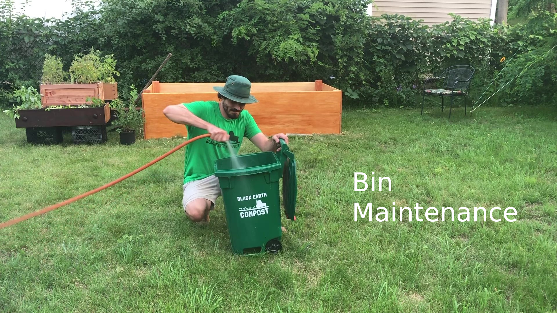 Man in green shirt and hat watering a green compost bin labeled Black Earth Compost in a grassy backyard.