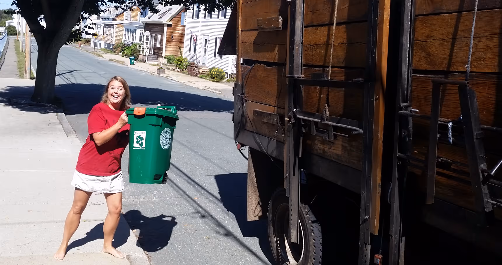 Woman in a red shirt and white skirt happily holding a green recycling bin next to a wooden recycling truck on a sunny residential street.