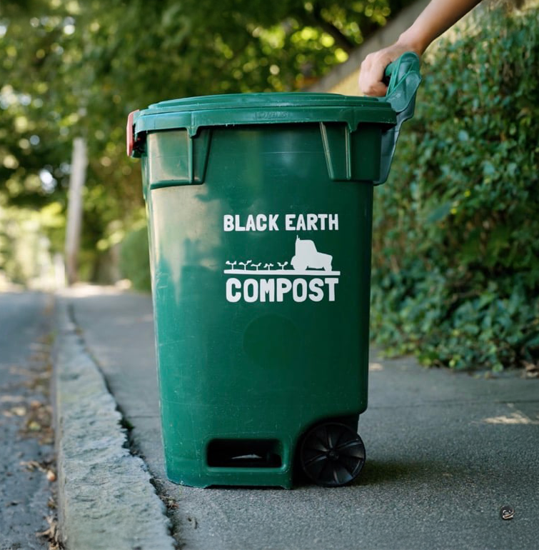 Green compost bin labeled 'Black Earth Compost' on a sidewalk curb with a hand holding the lid.