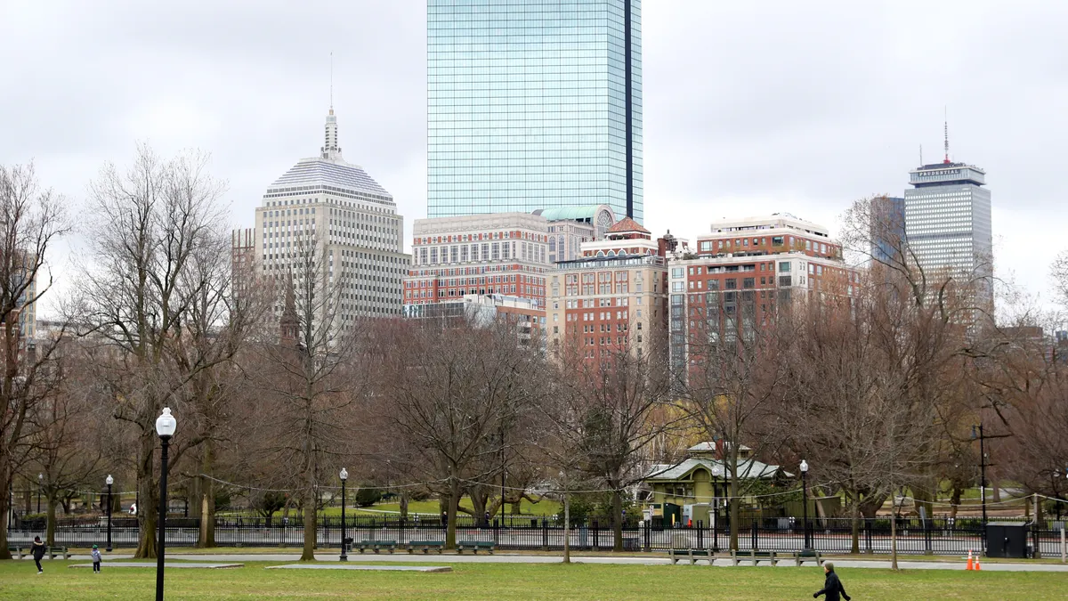 City park with leafless trees and green grass in front of tall modern and historic buildings on a cloudy day.
