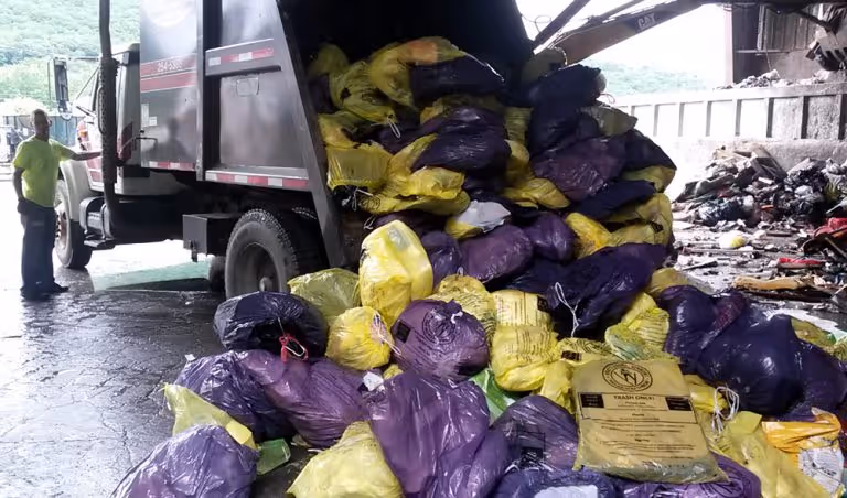 Man in neon green shirt standing beside a truck unloading purple and yellow bags filled with trash inside a waste management facility.
