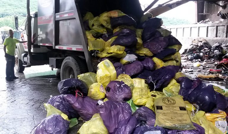 Man in neon green shirt standing beside a truck unloading purple and yellow bags filled with trash inside a waste management facility.