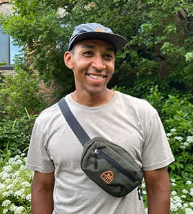 Smiling man wearing a gray cap, beige t-shirt, and green crossbody bag standing outdoors with green foliage and white flowers in the background.
