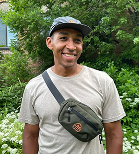 Smiling man wearing a gray cap, beige t-shirt, and green crossbody bag standing outdoors with green foliage and white flowers in the background.