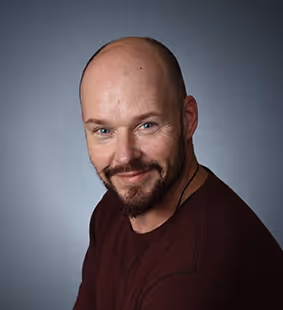 Smiling bald man with a beard wearing a dark red shirt against a gray background.