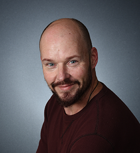 Smiling bald man with a beard wearing a dark red shirt against a gray background.