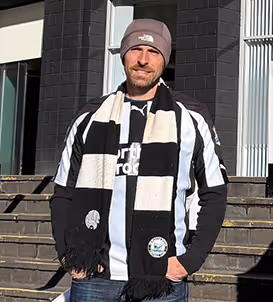 Man standing on outdoor steps wearing a gray beanie, black and white striped scarf, and black and white shirt under sunlight.