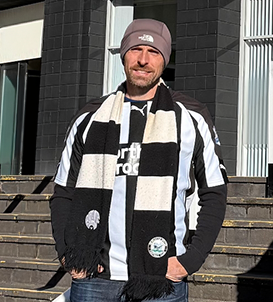 Man standing on outdoor steps wearing a gray beanie, black and white striped scarf, and black and white shirt under sunlight.