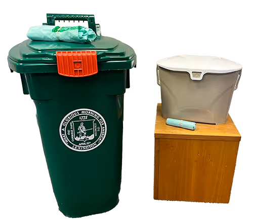 Green trash bin with a red latch and a printed city emblem, next to a wooden stool with a gray plastic bin and a rolled plastic bag on top.
