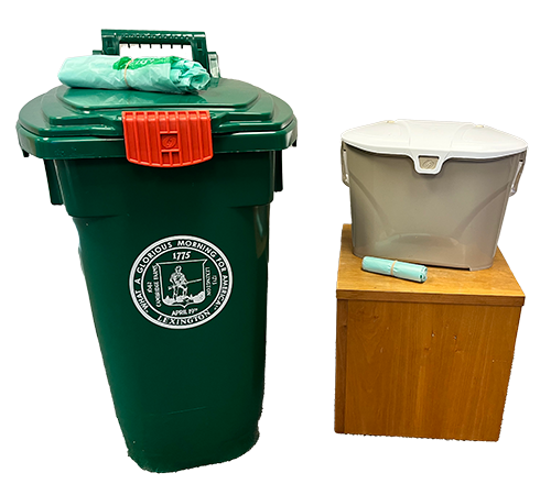 Green trash bin with a red latch and a printed city emblem, next to a wooden stool with a gray plastic bin and a rolled plastic bag on top.