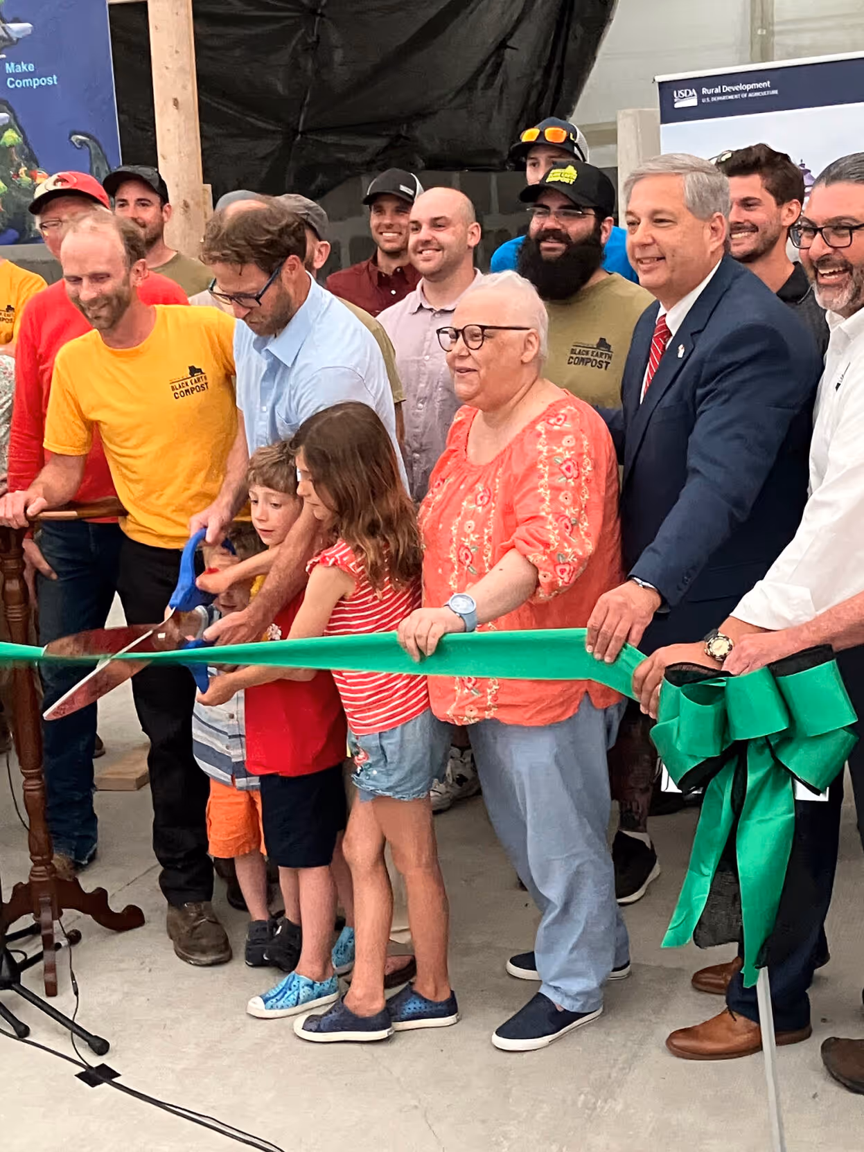 Group of people, including children and adults, cutting a green ribbon at a ceremony with smiles and casual to business attire.