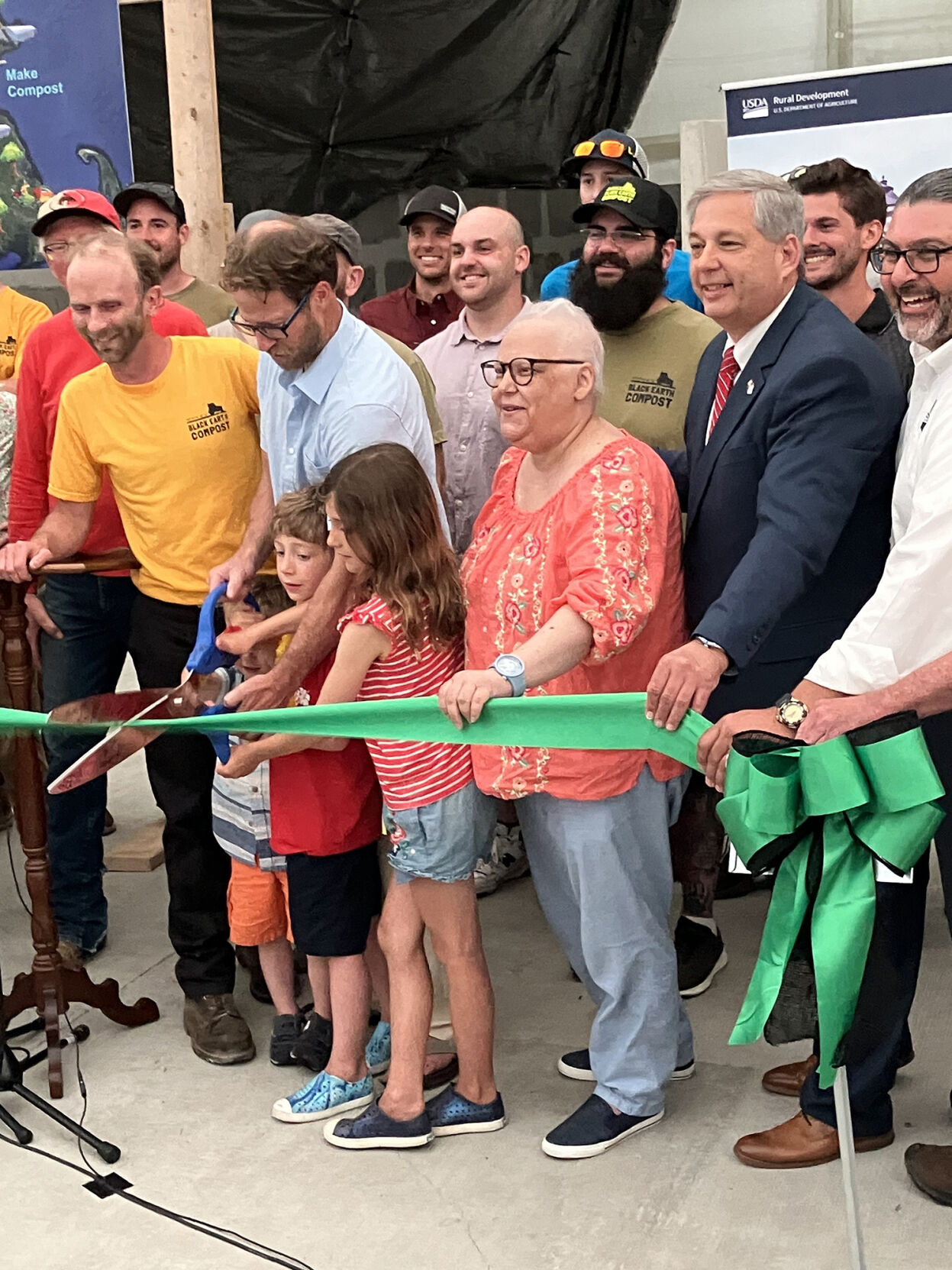 Group of people, including children and adults, cutting a green ribbon at a ceremony with smiles and casual to business attire.
