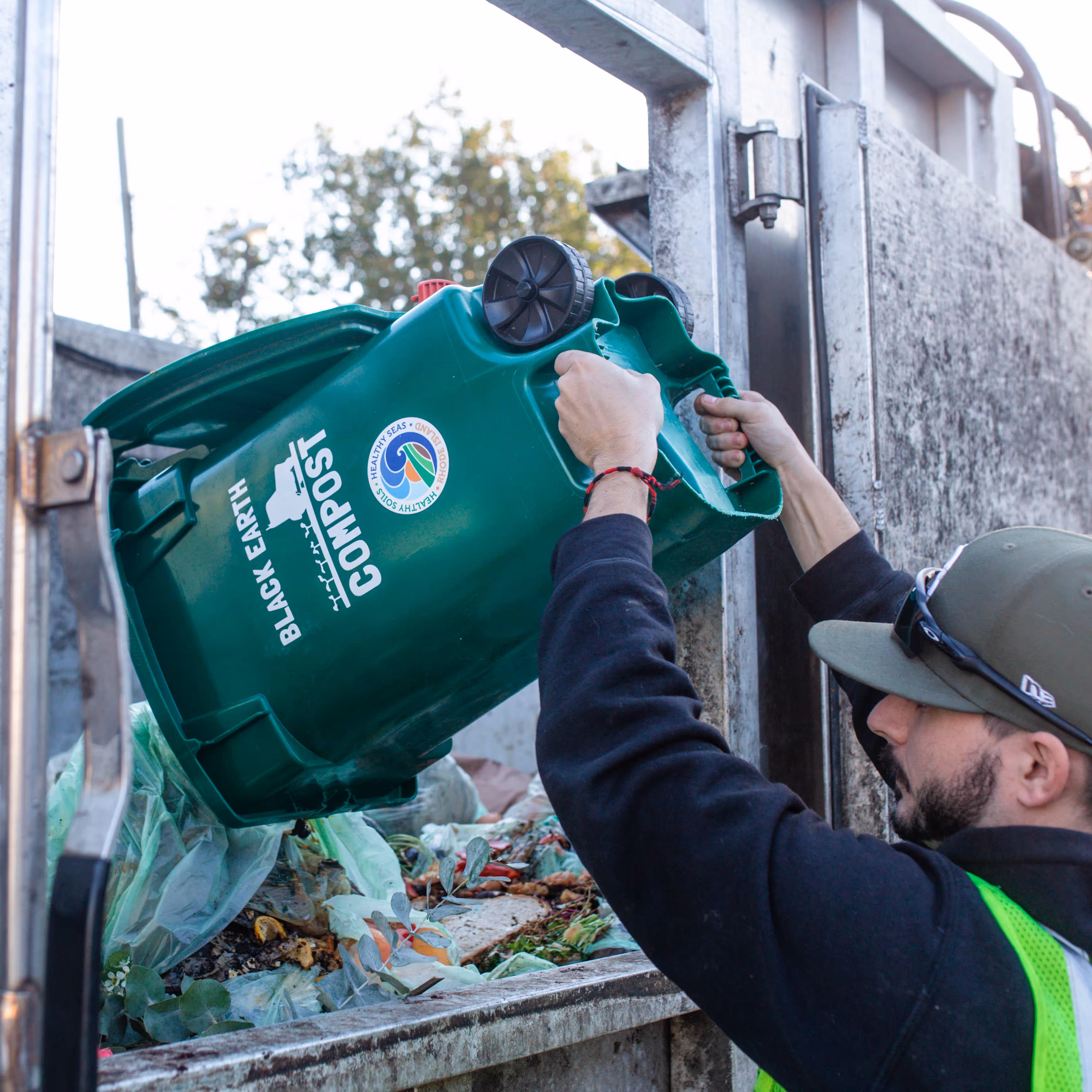 Person wearing a green cap and safety vest emptying a green compost bin labeled Black Earth into a garbage truck.