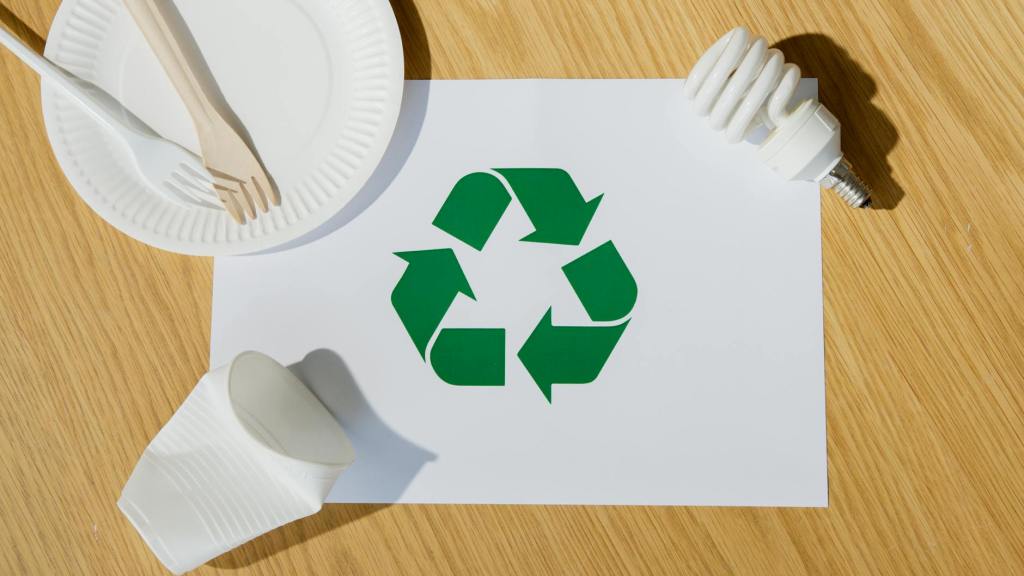 Green recycling symbol printed on white paper surrounded by biodegradable utensils on a paper plate, a broken white plastic cup, and an energy-saving light bulb on a wooden surface.