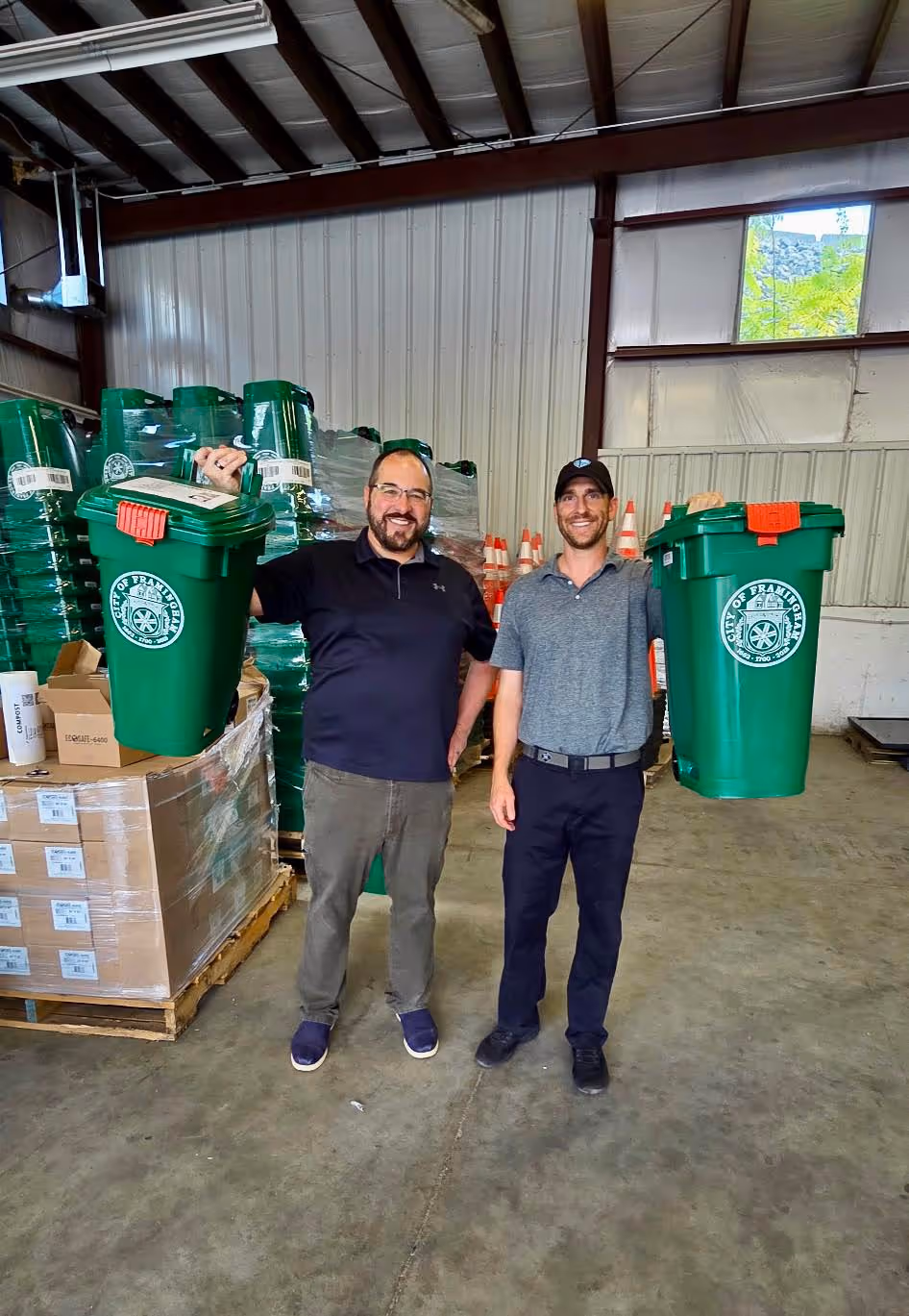Two men standing in a warehouse holding up green recycling bins labeled with the City of Framingham seal.