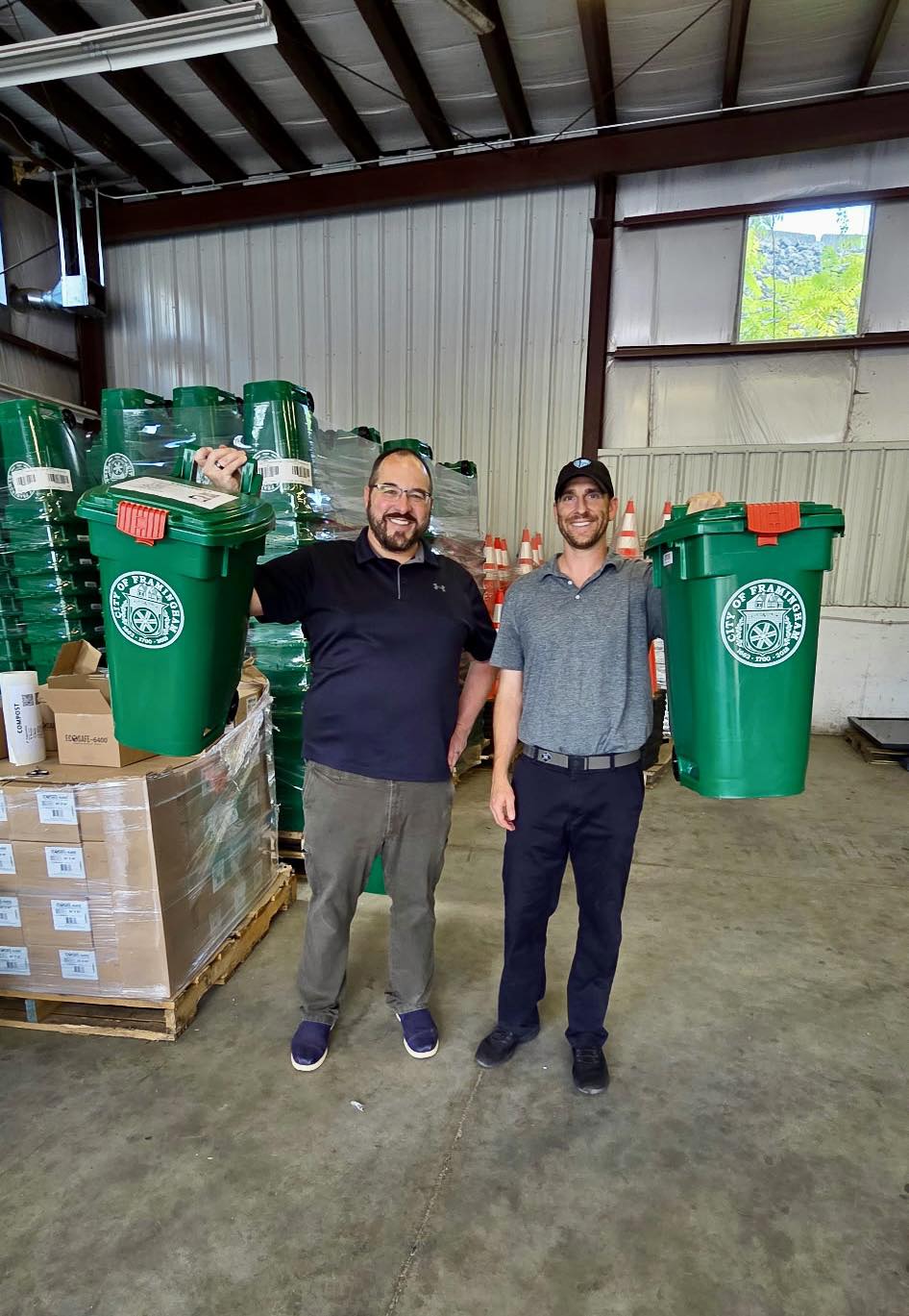 Two men standing in a warehouse holding up green recycling bins labeled with the City of Framingham seal.