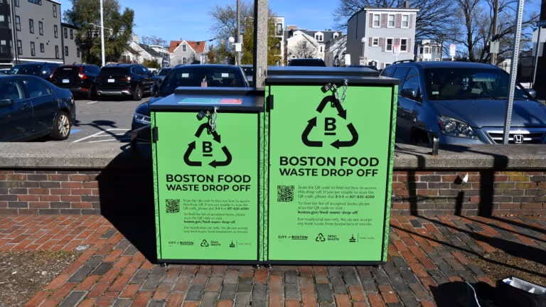 Two bright green Boston Food Waste Drop Off bins with locks, set on a brick sidewalk with parked cars and houses in the background.