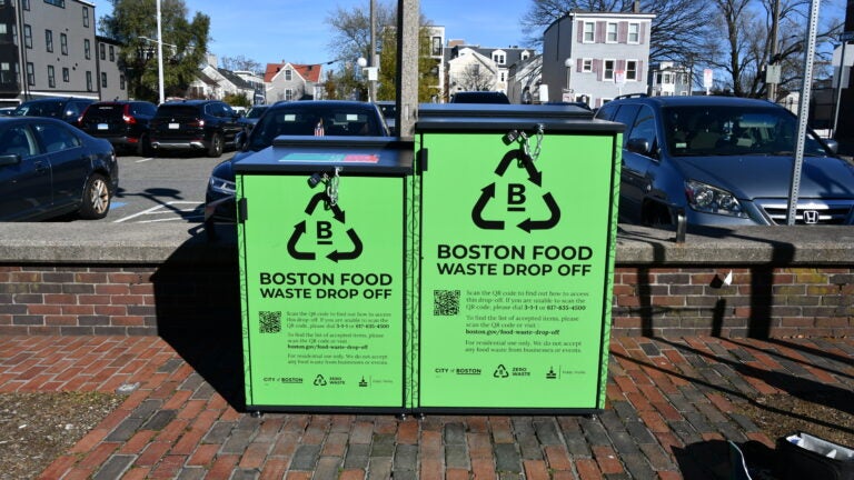 Two bright green Boston Food Waste Drop Off bins with locks, set on a brick sidewalk with parked cars and houses in the background.