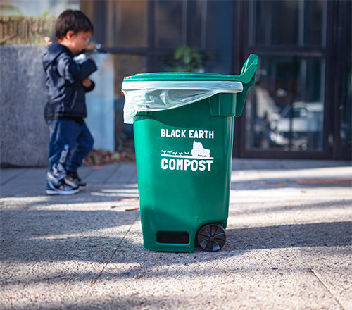 Green compost bin labeled 'BLACK EARTH COMPOST' with a child standing in the background.